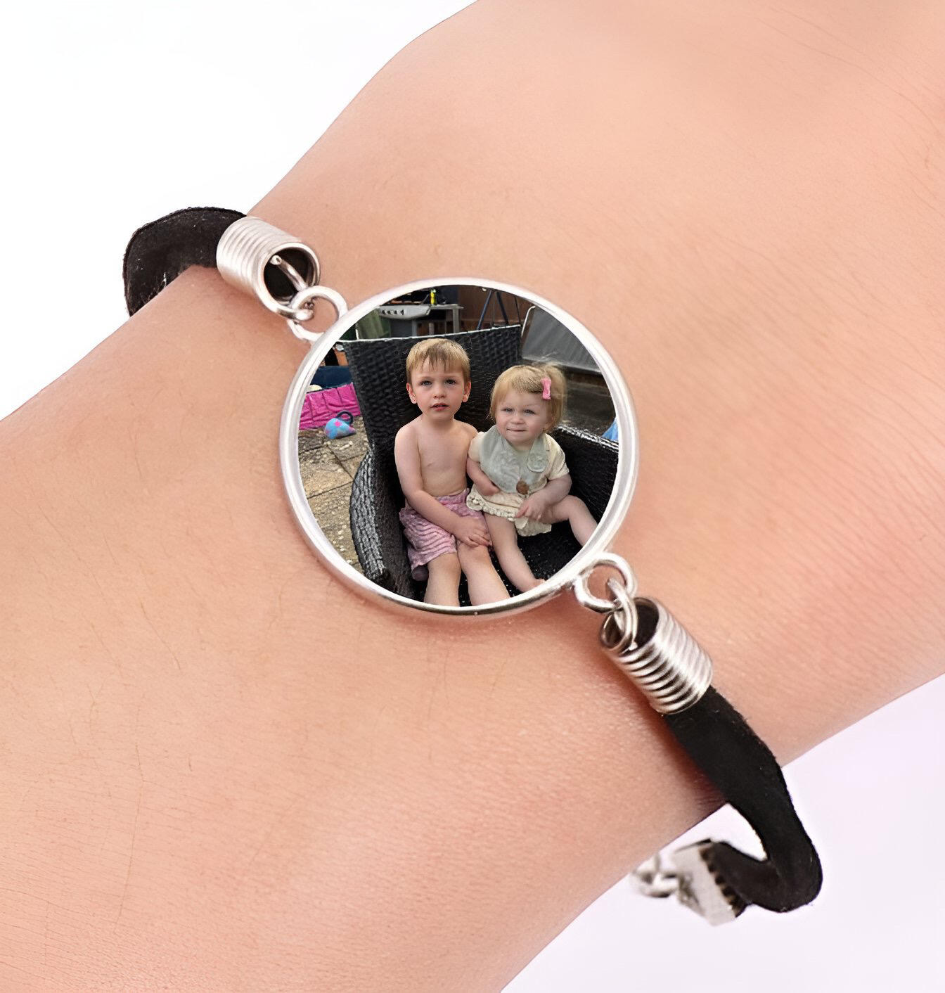 A circular silver charm bracelet with a black cord strap featuring a custom family photo of two young children sitting outside by QuestAbstract